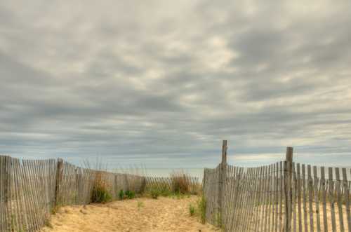 A sandy path leads through wooden fencing to a beach under a cloudy sky. Grassy plants peek through the sand.