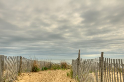 A sandy path leads through wooden fencing to a beach under a cloudy sky. Grassy plants peek through the sand.