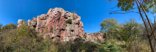 Panoramic view of a rocky cliff surrounded by greenery under a clear blue sky.
