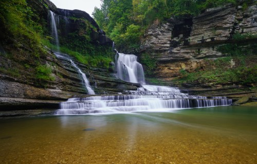 A serene waterfall cascades over rocky cliffs into a clear pool, surrounded by lush greenery.