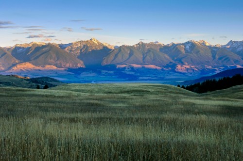 A scenic view of rolling hills and mountains under a clear blue sky, with sunlight illuminating the peaks.