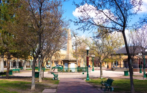 A park with trees, benches, and a monument under a blue sky with clouds, featuring a path leading through the area.