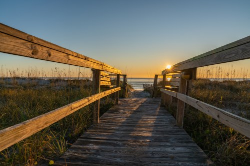 A wooden walkway leads to a beach at sunset, with tall grass on either side and the sun setting over the horizon.