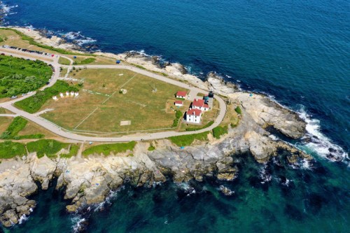 Aerial view of a coastal area with a lighthouse, grassy land, and rocky shoreline beside blue ocean waters.