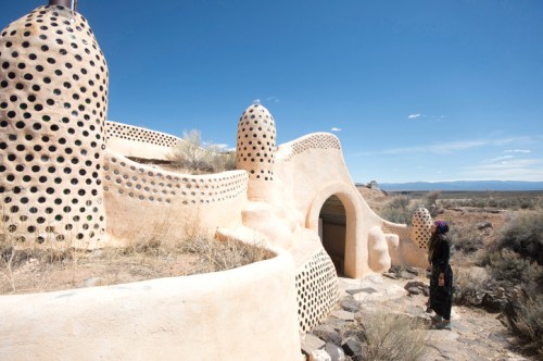 A person stands near a unique, organic-shaped building with dotted walls against a clear blue sky and desert landscape.