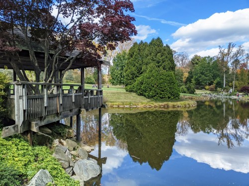 A serene pond reflects a gazebo and trees under a blue sky with fluffy clouds, surrounded by lush greenery.