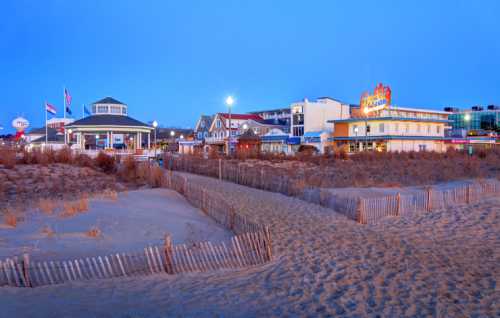 A beach scene at dusk featuring sand dunes, wooden fences, and colorful buildings along the shoreline.