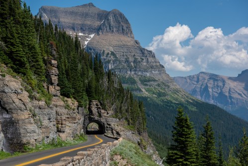 A scenic mountain road winds through lush greenery, with a tunnel and towering peaks under a blue sky with clouds.