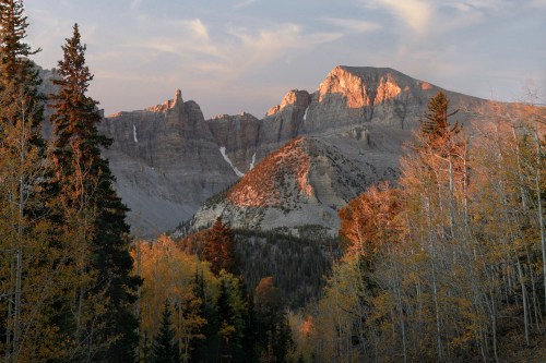 Sunlit mountains rise above a forest of autumn trees, with a clear sky and soft clouds in the background.