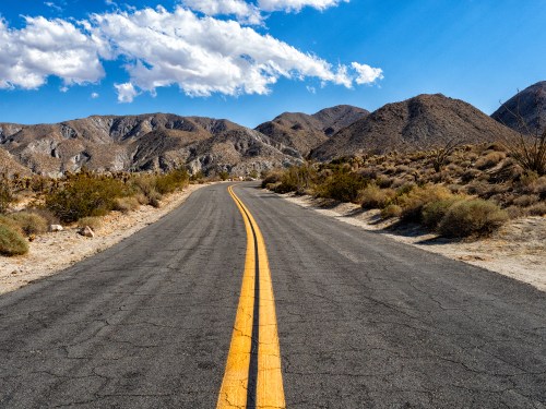 A winding road stretches through a desert landscape, flanked by mountains and dotted with sparse vegetation under a blue sky.