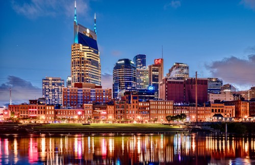 Nashville skyline at dusk, featuring illuminated buildings reflecting in the water.