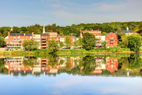 A serene riverside view of colorful buildings reflecting in calm water, surrounded by lush green trees.