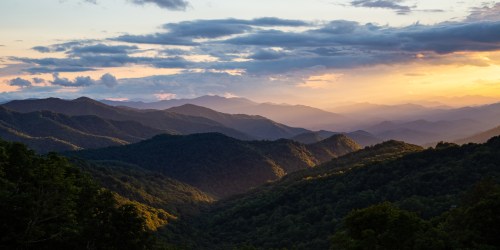 A scenic view of rolling mountains at sunset, with layers of green hills and a colorful sky filled with clouds.