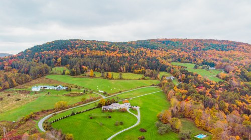 Aerial view of a scenic landscape with colorful autumn foliage, rolling hills, and a few houses along winding roads.