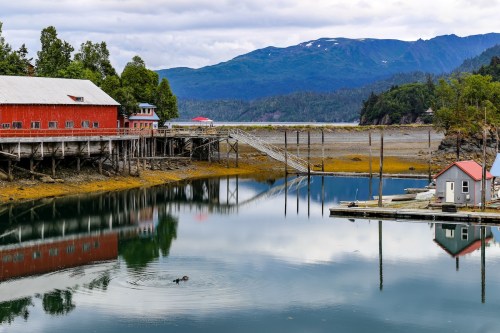 A red building on stilts by a calm waterway, surrounded by mountains and trees, with a small boat docked nearby.