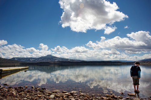 A boy stands at the edge of a calm lake, reflecting clouds and mountains under a bright blue sky.