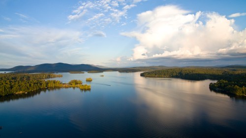 A serene lake surrounded by lush greenery and distant mountains under a blue sky with scattered clouds.