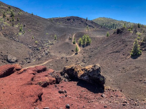 A rugged volcanic landscape with a winding dirt path, surrounded by dark lava rock and sparse greenery under a clear blue sky.