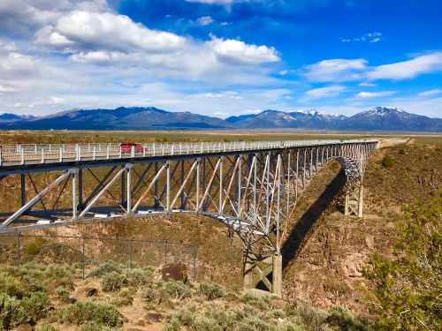 A red truck crosses a tall metal bridge over a canyon, with mountains and blue skies in the background.
