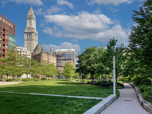 A city park with green grass and trees, featuring a path and a historic clock tower in the background under a blue sky.