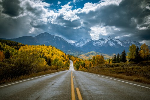 A winding road leads through vibrant autumn trees, with snow-capped mountains and dramatic clouds in the background.