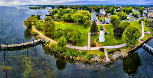 Aerial view of a park with a lighthouse, pathways, and trees by a calm waterfront under a cloudy sky.