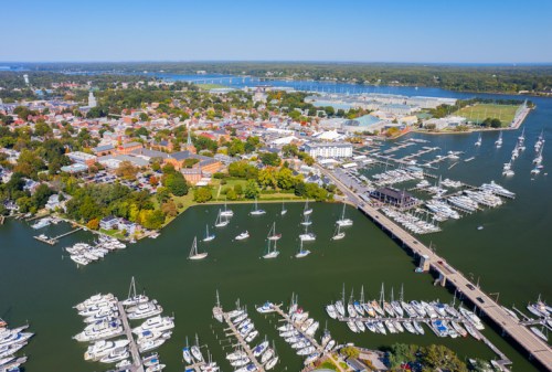 Aerial view of a vibrant waterfront town with marinas, sailboats, and lush greenery under a clear blue sky.