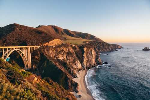 A scenic view of a coastal bridge over cliffs, with waves crashing on the shore and mountains in the background at sunset.