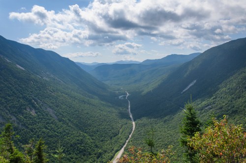 A scenic view of a lush green valley surrounded by mountains under a partly cloudy sky. A winding road cuts through the landscape.