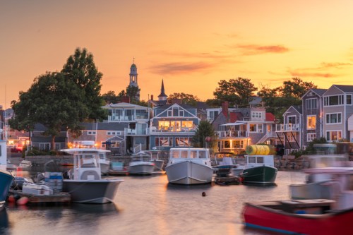 A scenic harbor at sunset, featuring boats and charming waterfront homes with trees and a church steeple in the background.