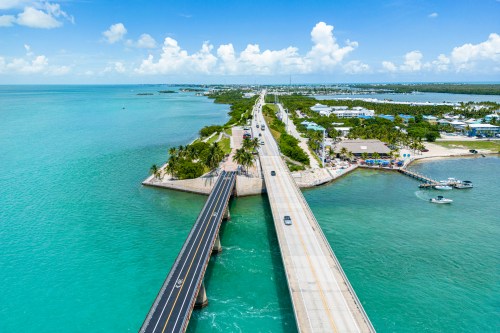Aerial view of a coastal road with palm trees, water, and boats under a clear blue sky.