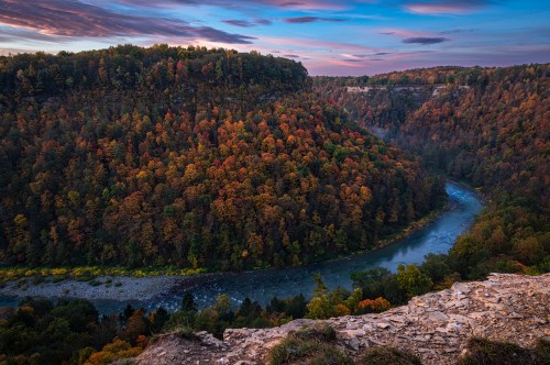 A scenic view of a winding river surrounded by vibrant autumn foliage and cliffs under a colorful sky.