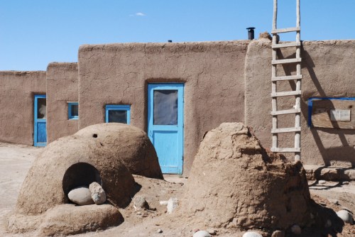 Adobe-style buildings with blue doors, a wooden ladder, and clay structures in a desert landscape.
