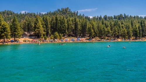 A serene lake surrounded by lush green trees, with people enjoying the beach and boating under a clear blue sky.
