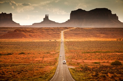 A long, empty road stretches through a desert landscape, flanked by towering rock formations under a cloudy sky.