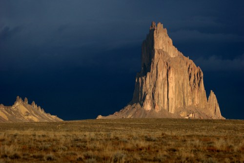 A dramatic rock formation rises against a dark sky, surrounded by a vast, open landscape.