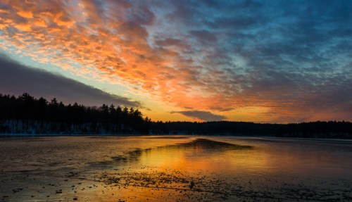 A serene lake at sunset, reflecting vibrant orange and blue hues in the sky, surrounded by trees.