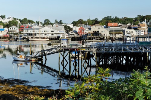 A calm harbor scene with a wooden pier, boats, and colorful buildings reflecting in the water.