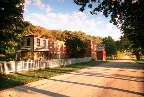 A peaceful rural scene featuring historic buildings along a tree-lined road under a blue sky with scattered clouds.