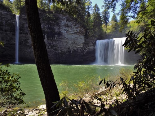 A serene landscape featuring two waterfalls cascading into a green pool, surrounded by lush trees and rocky cliffs.