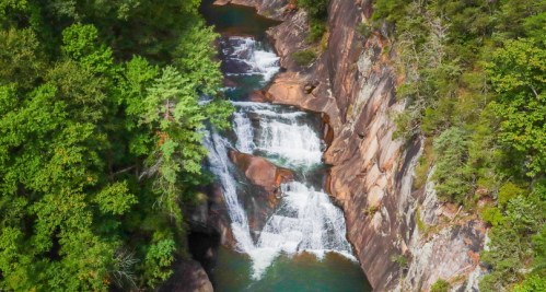 Aerial view of a cascading waterfall surrounded by lush green trees and rocky cliffs.