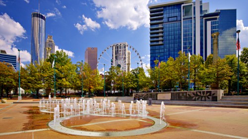 City park with a fountain, skyscrapers, and a Ferris wheel under a blue sky with fluffy clouds.