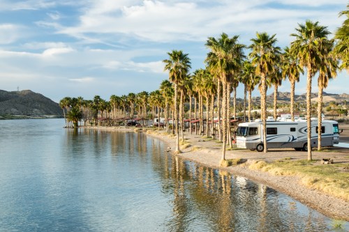 A serene lakeside scene with palm trees, an RV parked along the shore, and mountains in the background under a blue sky.
