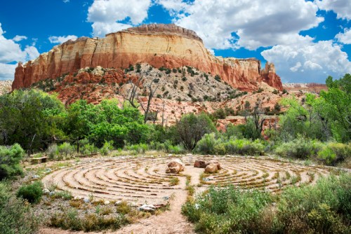 A serene labyrinth surrounded by lush greenery and dramatic red rock formations under a blue sky with clouds.