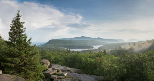 A panoramic view of lush green mountains and a serene lake under a blue sky with scattered clouds.