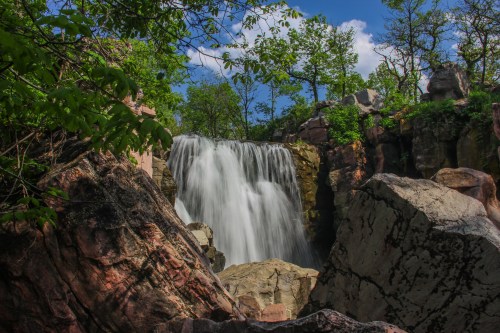 A serene waterfall cascades over rocks, surrounded by lush greenery and a bright blue sky.