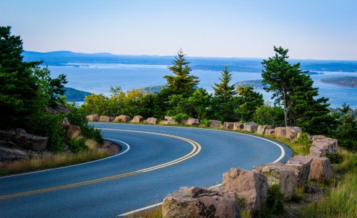 A winding road curves along a scenic overlook with trees and a view of a lake and distant hills.