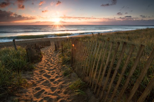 A sandy path leads to a beach at sunset, with gentle waves and clouds in the sky.