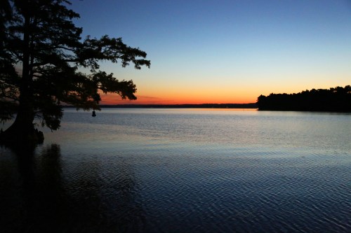 A serene lake at sunset, with calm waters reflecting the colorful sky and silhouettes of trees along the shore.