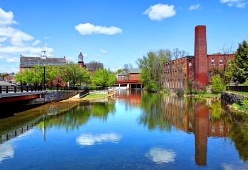 A serene river scene with reflections of trees and buildings under a blue sky with fluffy clouds.